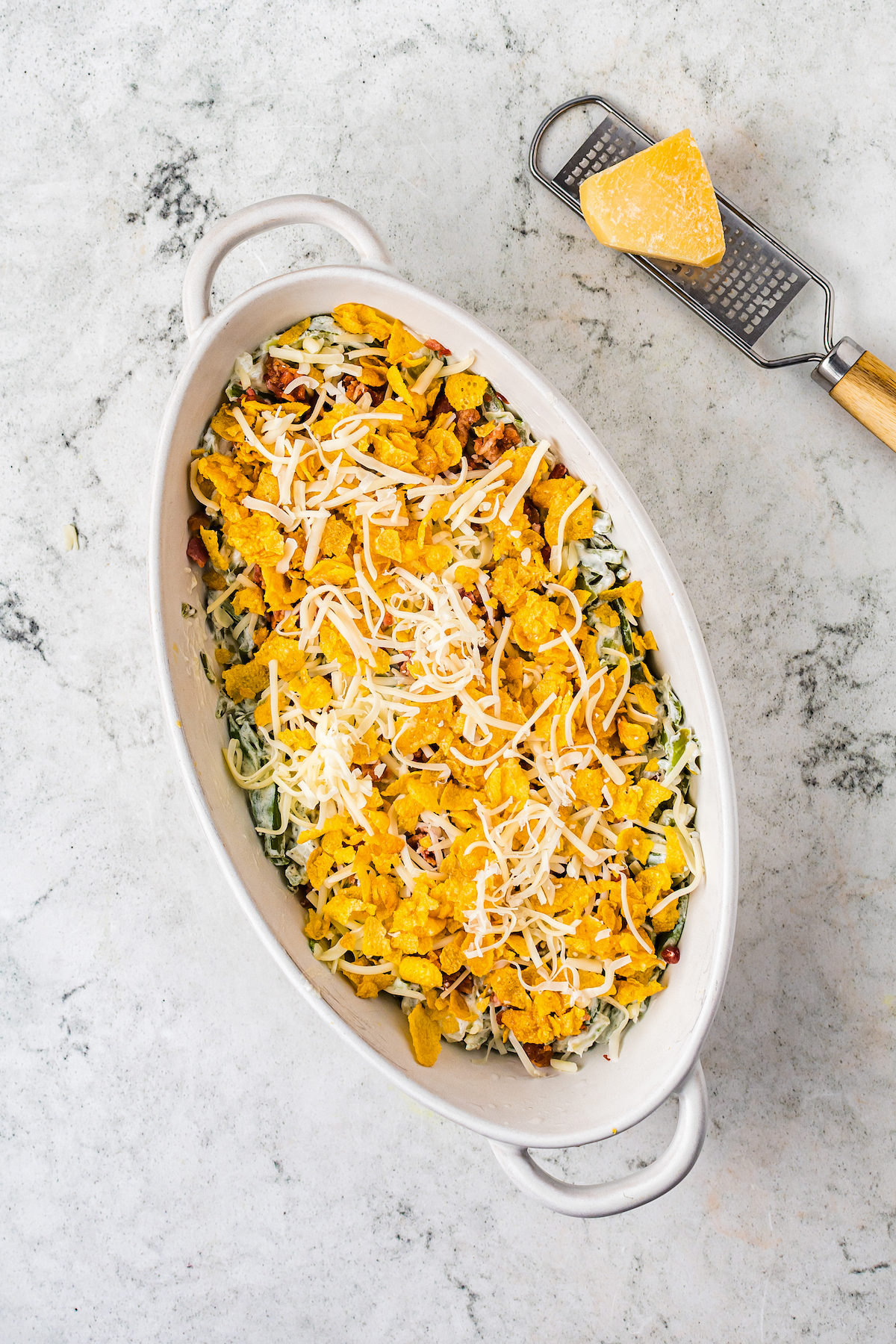 An unbaked casserole topped with green beans, cornflakes, and cheese.
