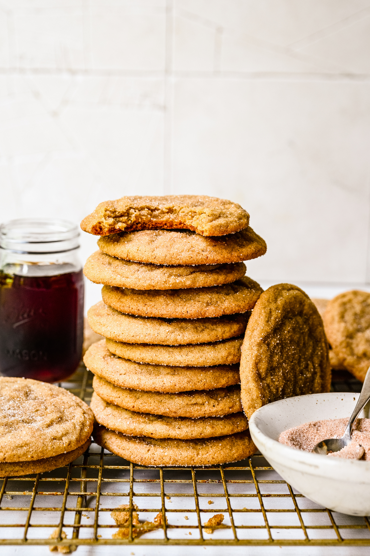 A stack of cookies. The cookie on top has had a bite taken from it, while another cookie is propped up against the stack. Maple syrup and cinnamon sugar are nearby on the wire rack.