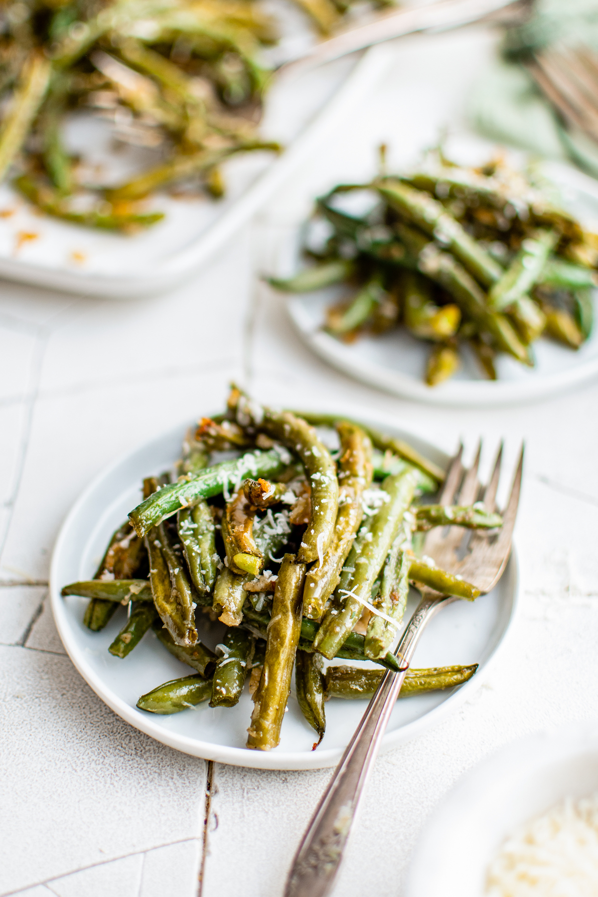 Two small white plates with servings of roasted green beans and forks. A larger platter of green beans is in the background of the shot.