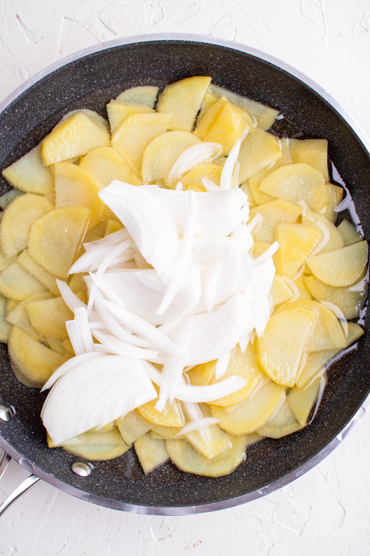A skillet filled with half-moon slices of potatoes in oil, with slices of white onion piled on top