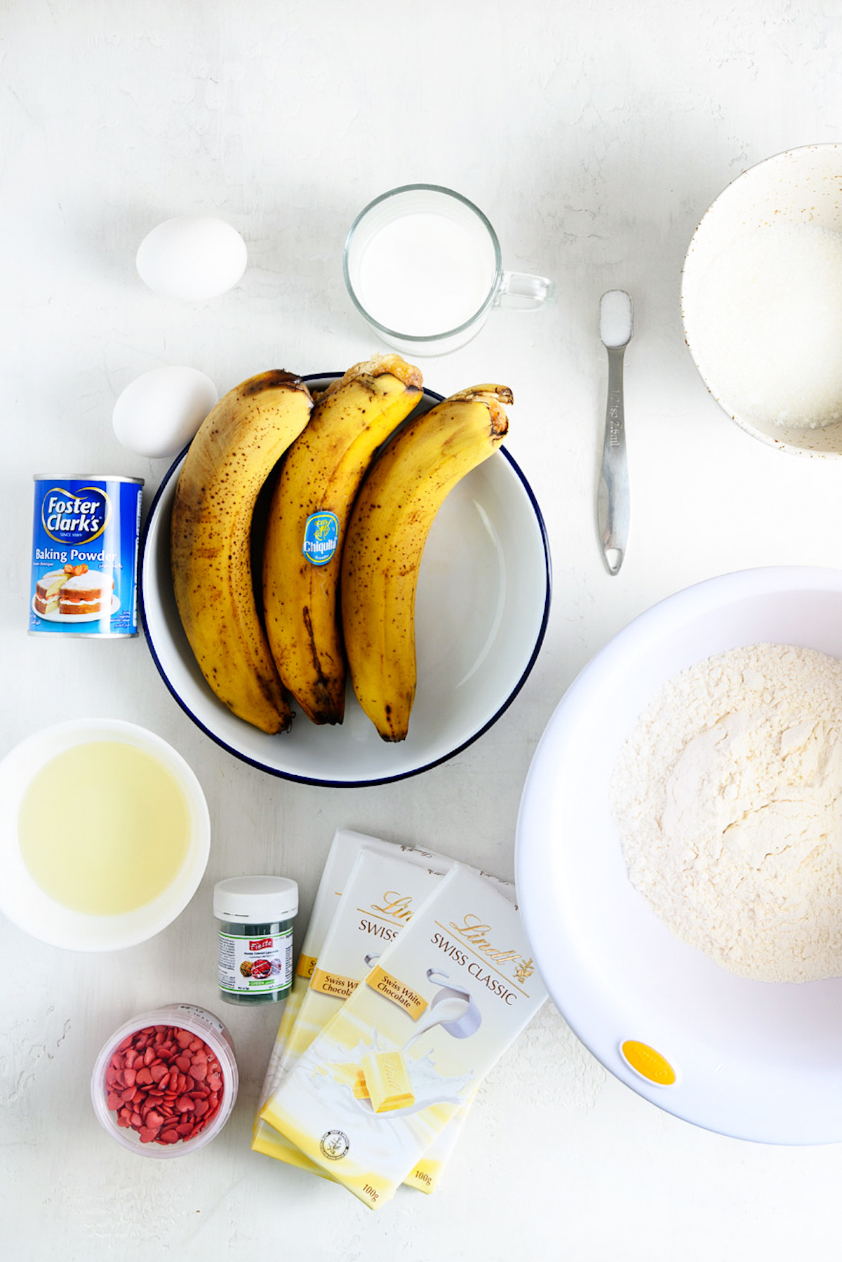 From top left, ingredients for banana cake recipe: Eggs, milk, salt, sugar, baking powder, bananas, flour, vegetable oil, sprinkles, food coloring, white chocolate.