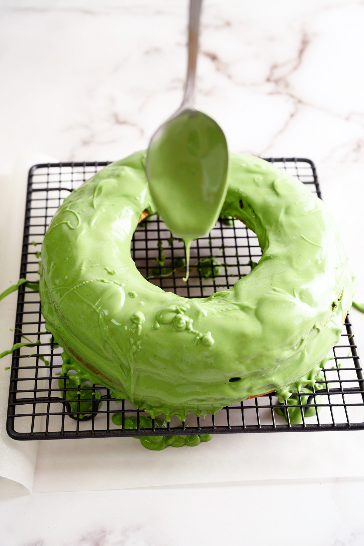 Melted white chocolate tinted green, being poured over a cake on a wire rack.