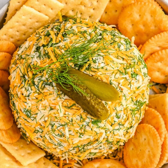 A dill pickle cheese ball, garnished with a gherkin, sprig of mint, and piles of crackers.