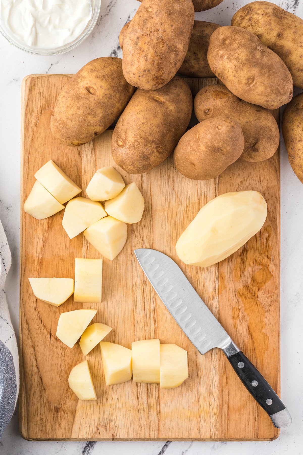 Overhead shot of a wooden cutting board. Two peeled potatoes are on the board with a knife. One has been cut into cubes. More potatoes are near the cutting board.