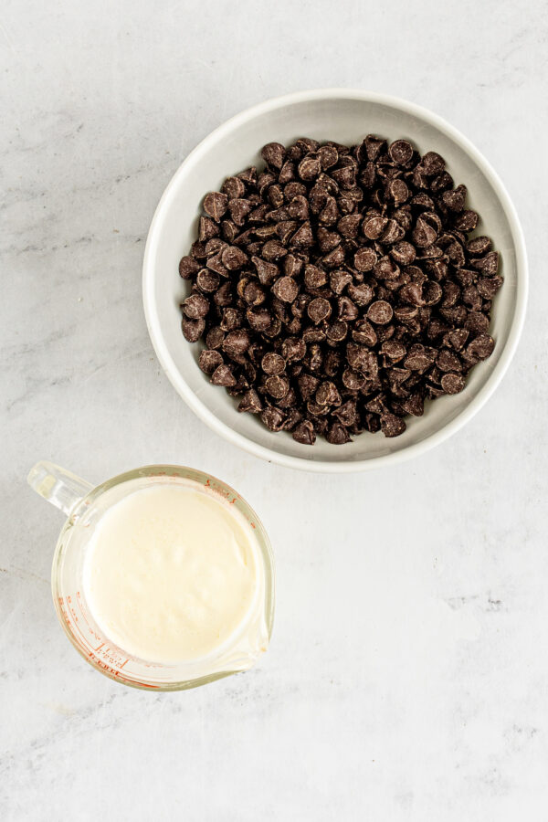 A bowl of semisweet chocolate chips next to a measuring cup of heavy cream.