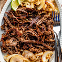 A serving plate with shredded crispy beef, sautéed onions, lime wedges and two forks.