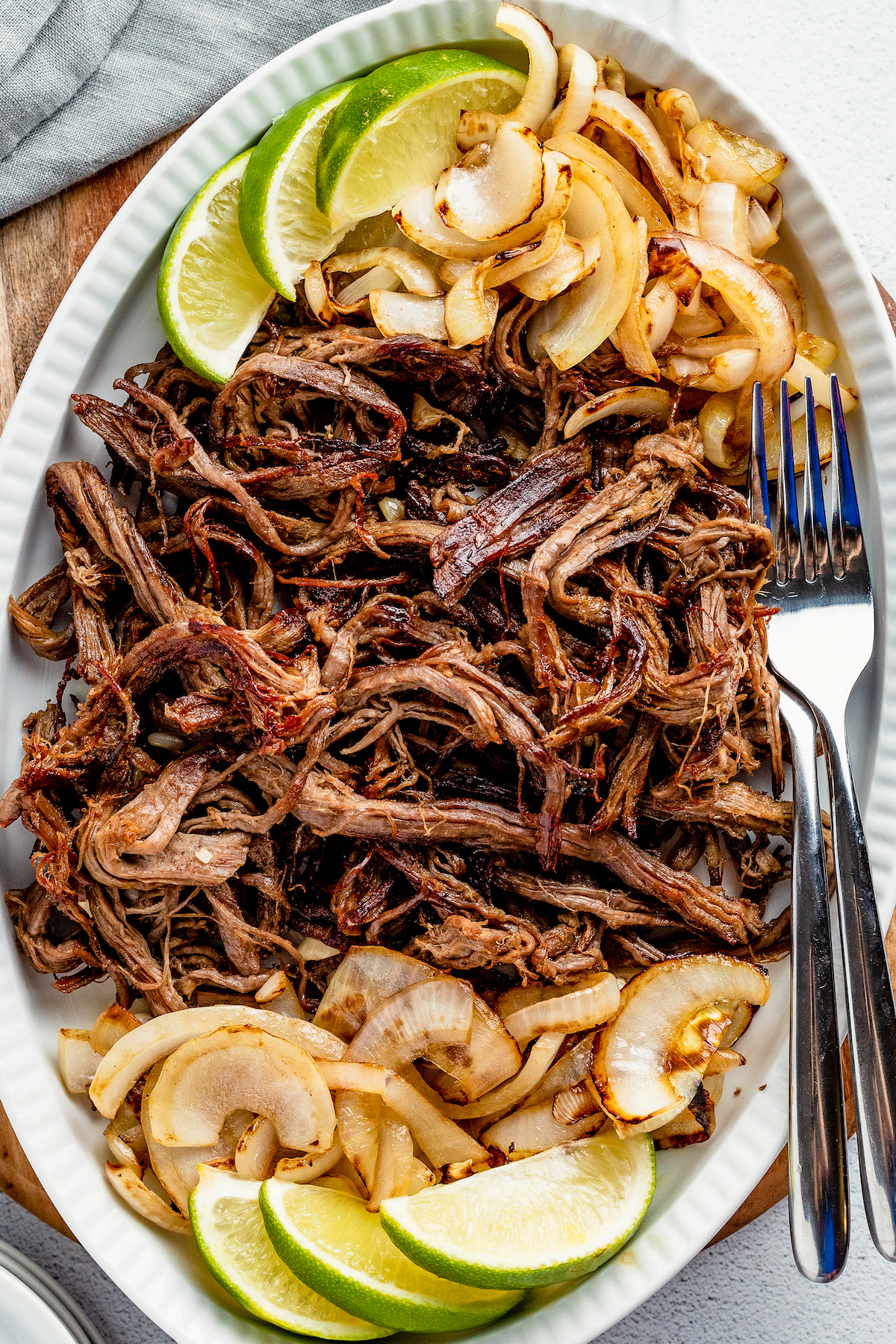 A serving plate with shredded crispy beef, sautéed onions, lime wedges and two forks.