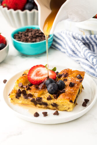 Slice of chocolate chip croissant French toast bake with blueberries, a strawberry, and chocolate chips on top. Honey is being drizzled over it.