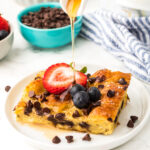A square piece of chocolate chip bread pudding, reminiscent of a decadent French toast casserole, is topped with sliced strawberries and blueberries on a white plate as syrup is poured over it. Bowls of berries and chocolate chips fill the background.