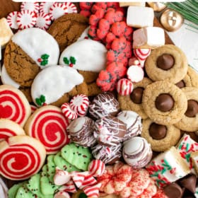 A tray filled with 7 types of christmas cookies and candies with bells and peppermint candies around the tray.