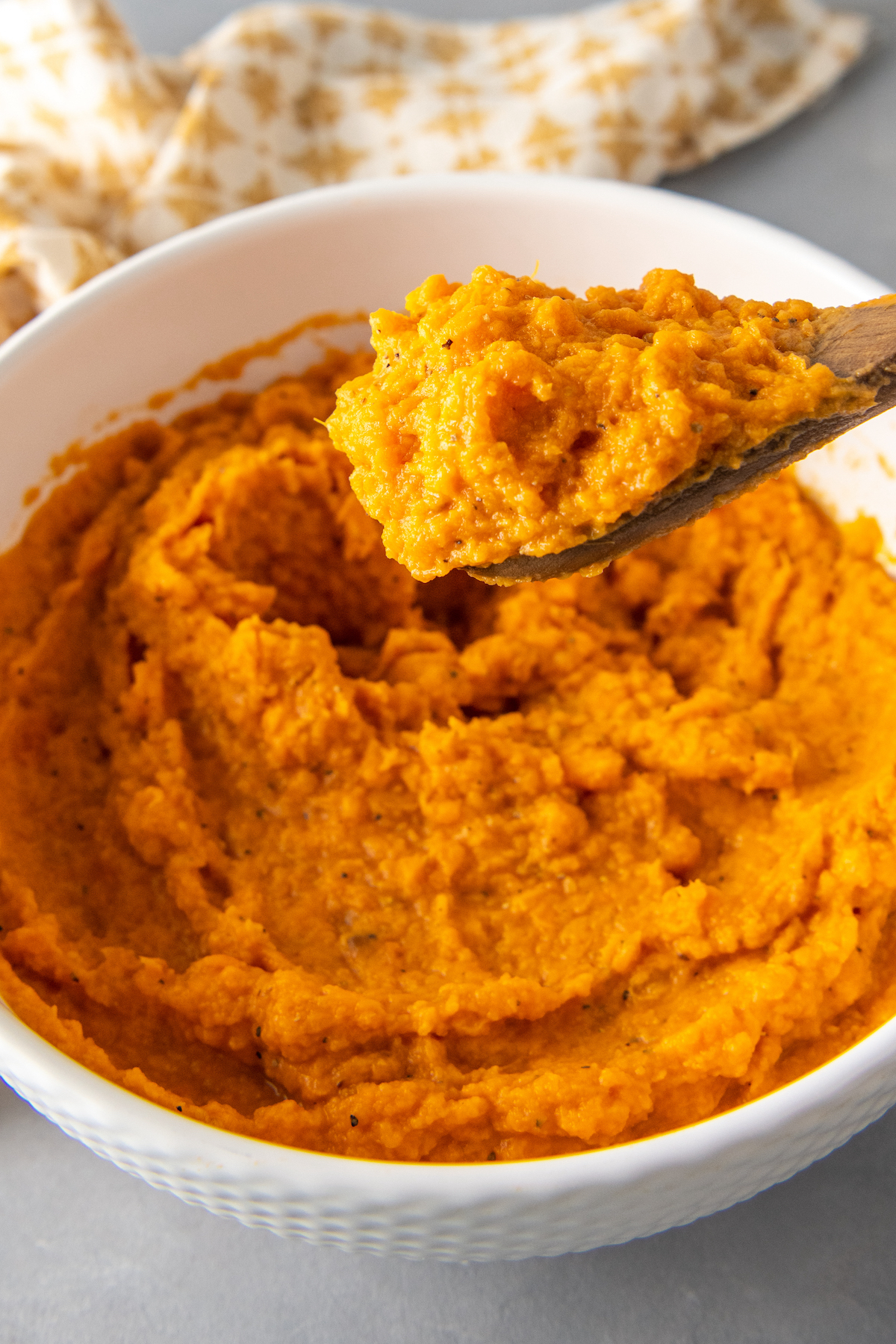 Mashed sweet potatoes in a bowl with seasonings with a wooden spoon scooping up some sweet potatoes.