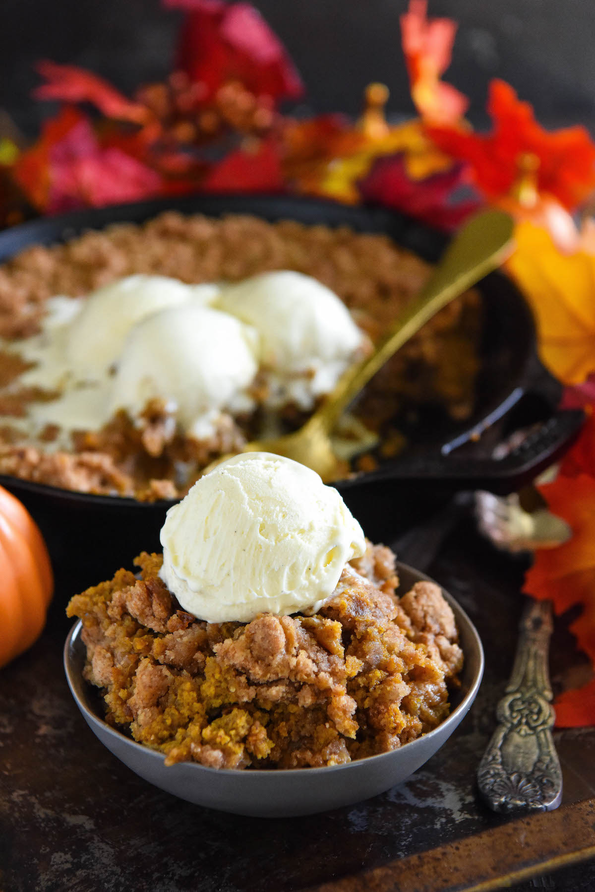 A cast iron skillet filled with pumpkin crisp and a serving in a bowl with a scoop of ice cream on top.