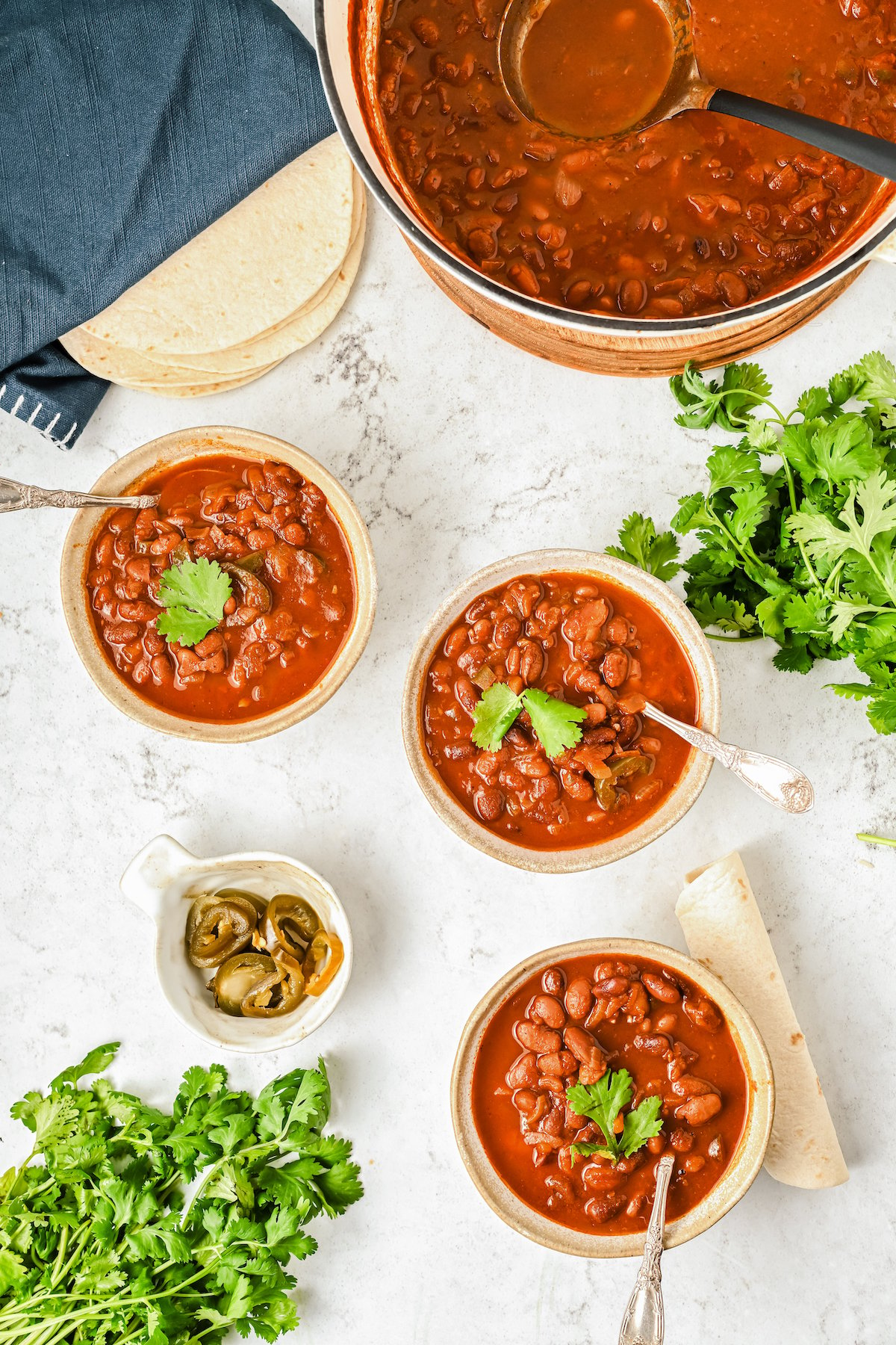 Three bowls of ranch-style beans on the table with cilantro and corn tortillas. The pot is on the side.