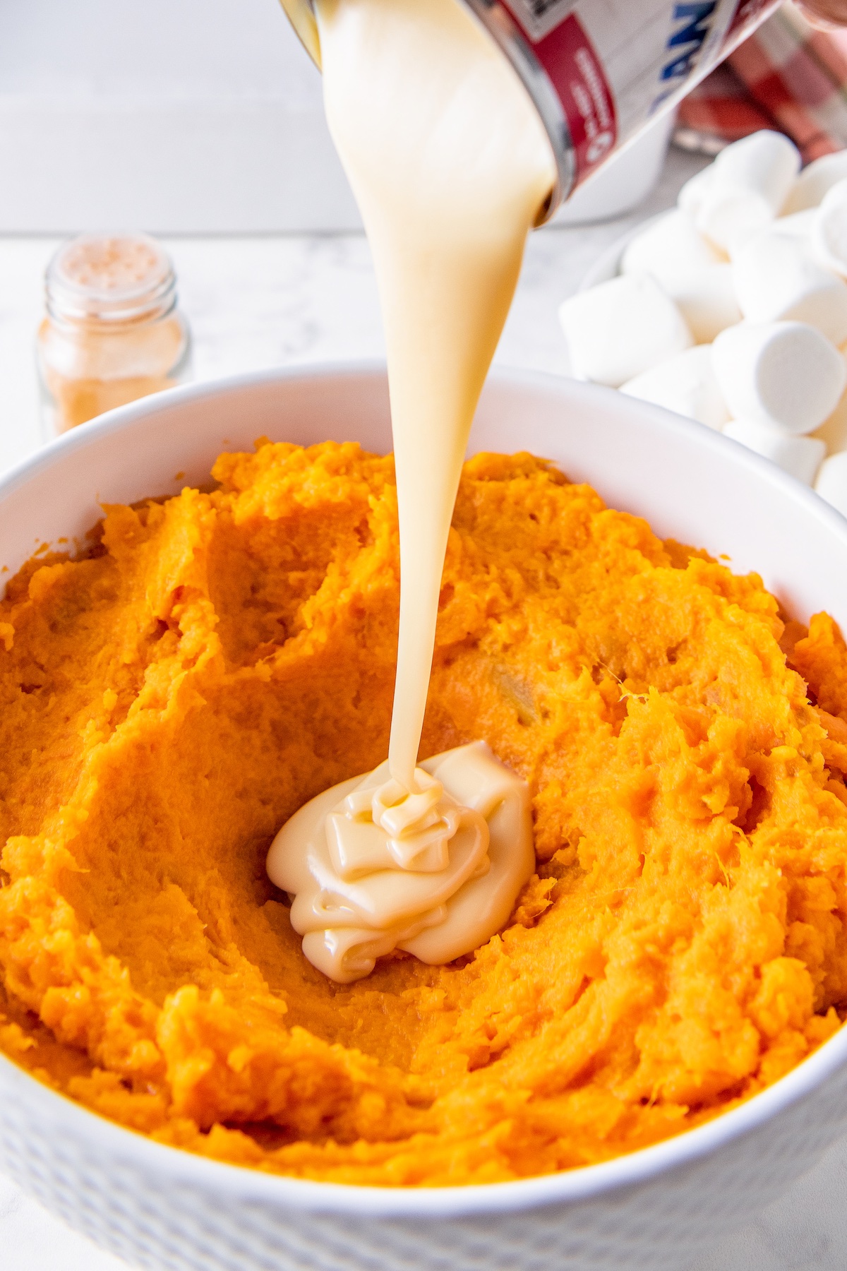 Sweetened condensed milk being poured into a bowl of mashed sweet potatoes.