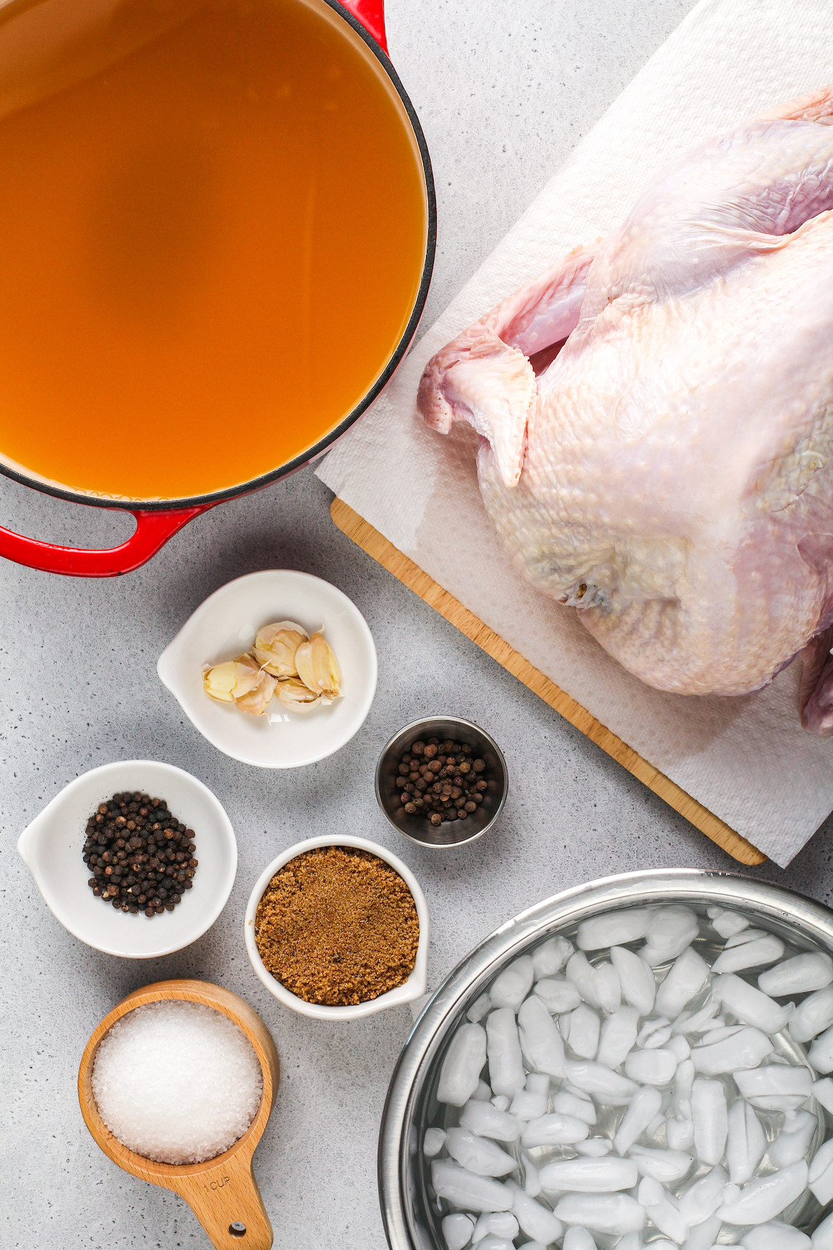 Ingredients for turkey brine recipe from top left: Vegetable stock, a whole raw turkey, garlic cloves, black peppercorns, allspice berries, dark brown sugar, kosher salt, ice water.