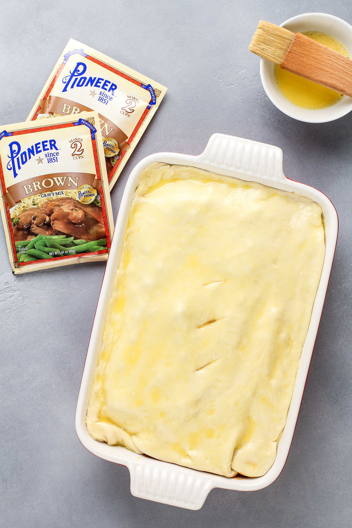 Puff pastry topped casserole brushed with butter and a bowl of butter with a pastry brush.