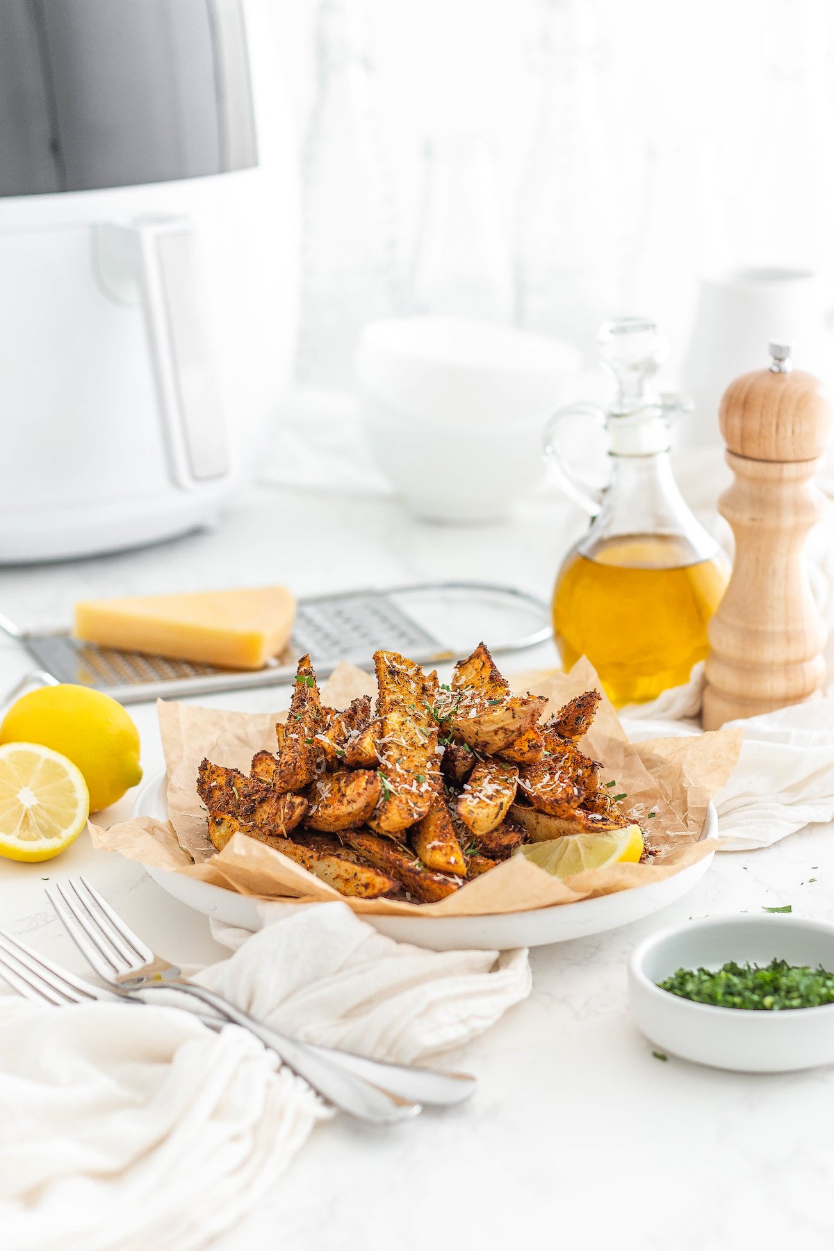 A table with a plate of seasoned potato wedges, an air fryer, a cloth napkin, and other items.