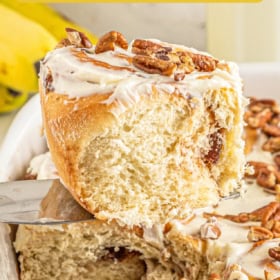 A banana bread cinnamon roll being lifted out of a tray of banana cinnamon rolls.