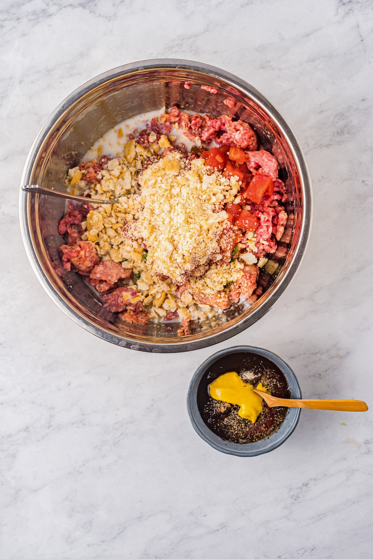 Raw hamburger, cracker crumbs. diced tomatoes, and other ingredients in a mixing bowl. A smaller bowl of topping ingredients is nearby on the work surface.
