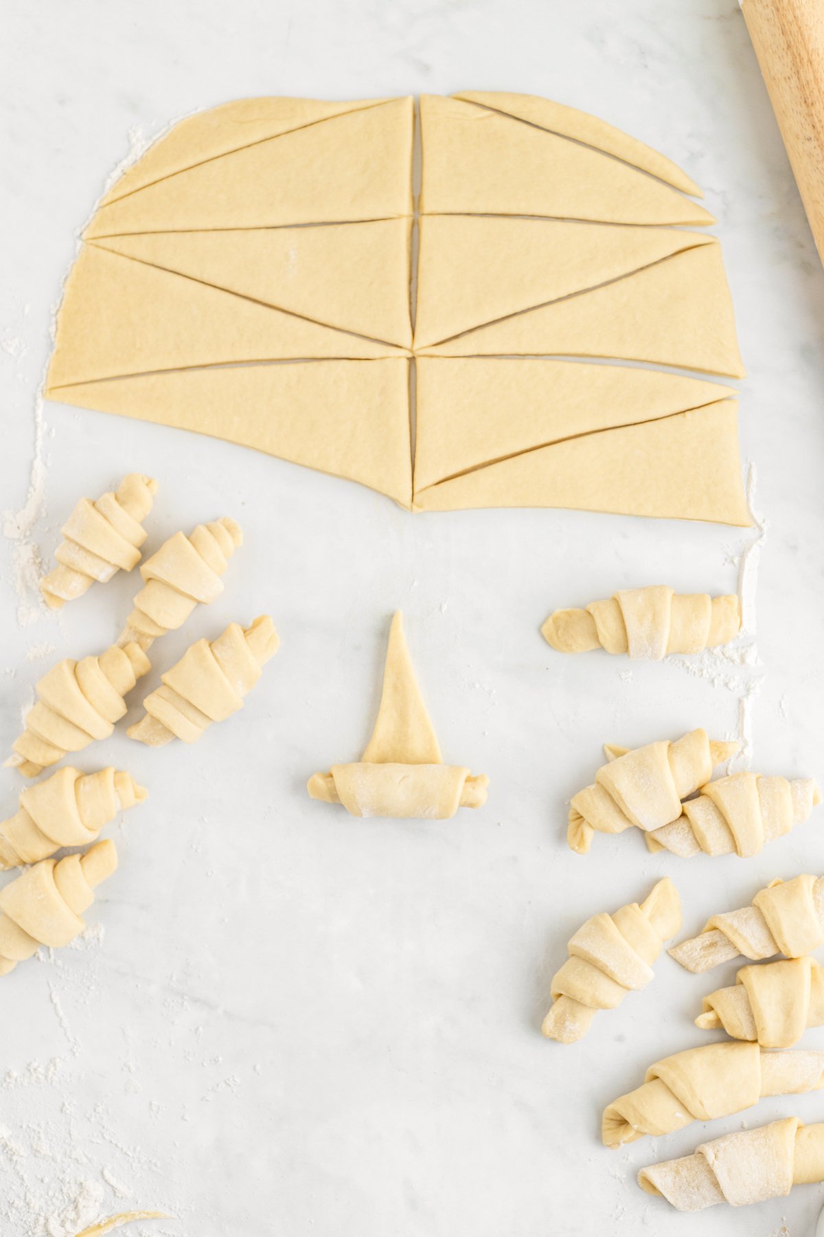 A half-rolled crescent made of dough. Other dough crescents and unrolled dough triangles are on the work surface as well.