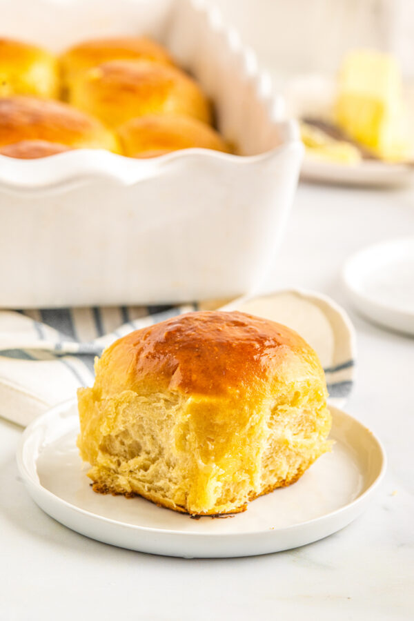 A soft, tender and sweet Hawaiian roll on a small white plate, with the pan of rolls and a dish of butter in the background of the shot.
