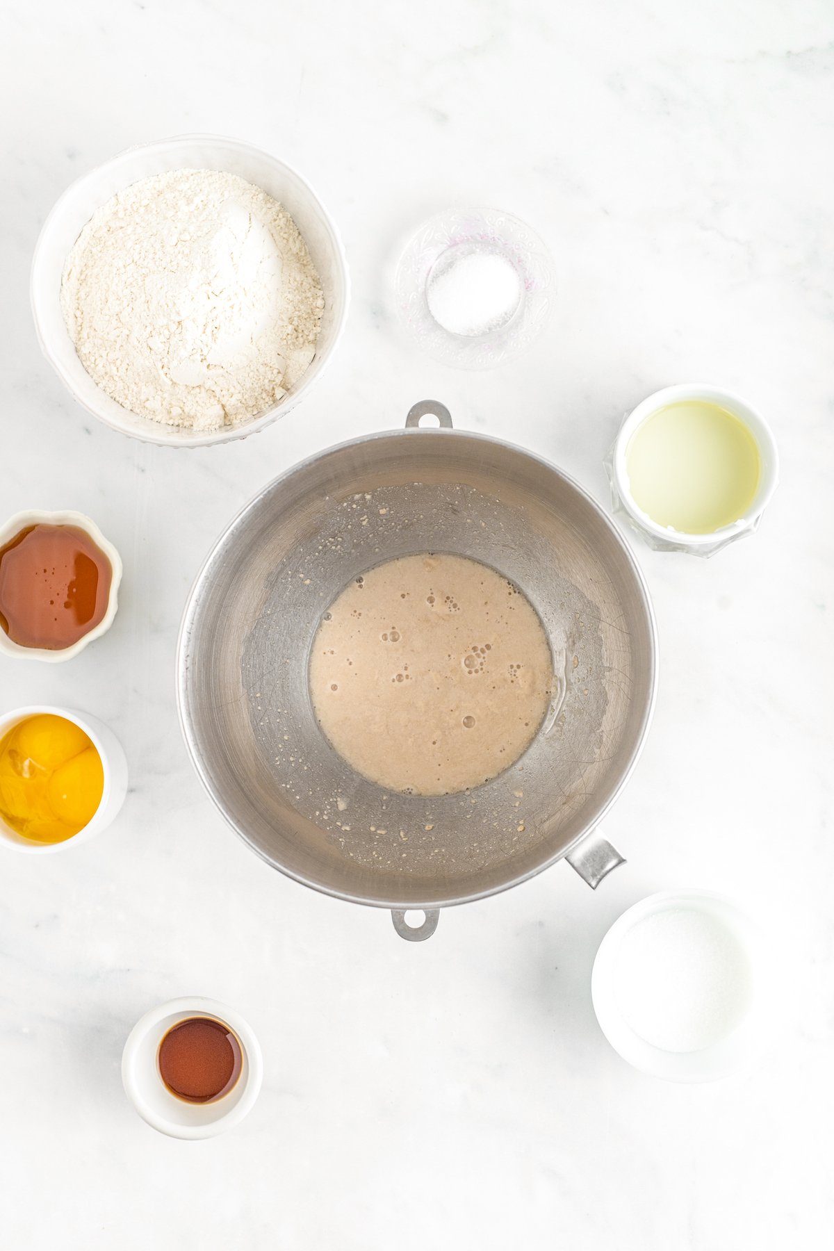 Yeast proving in a mixing bowl, with dishes of other ingredients arranged around the mixing bowl on the table.