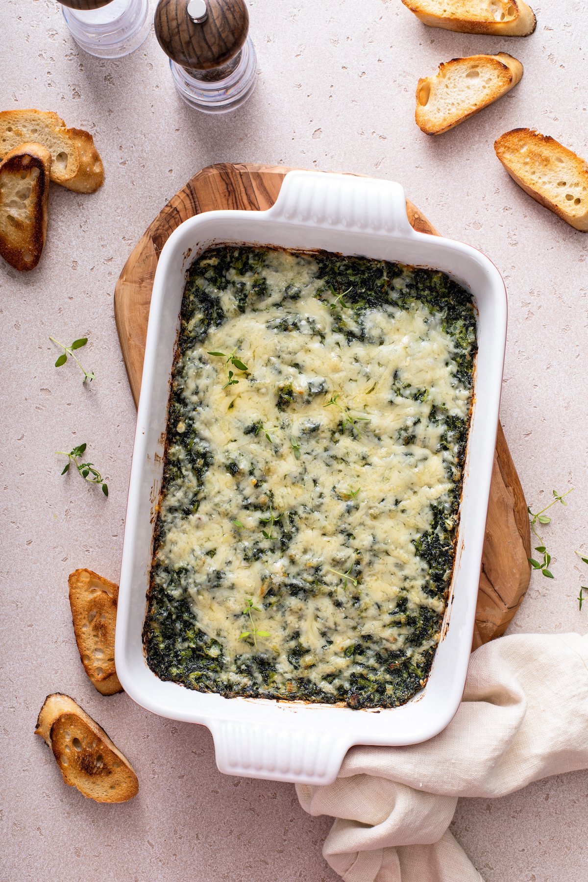 A baking dish filled with cheesy spinach casserole on a cutting board with a tea towel.