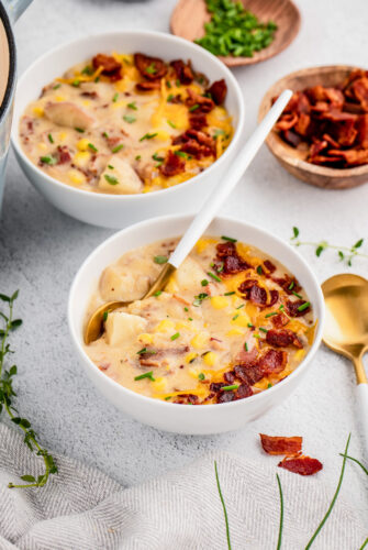 Two bowls of corn chowder with bacon, cheese and chives on top with a spoon.