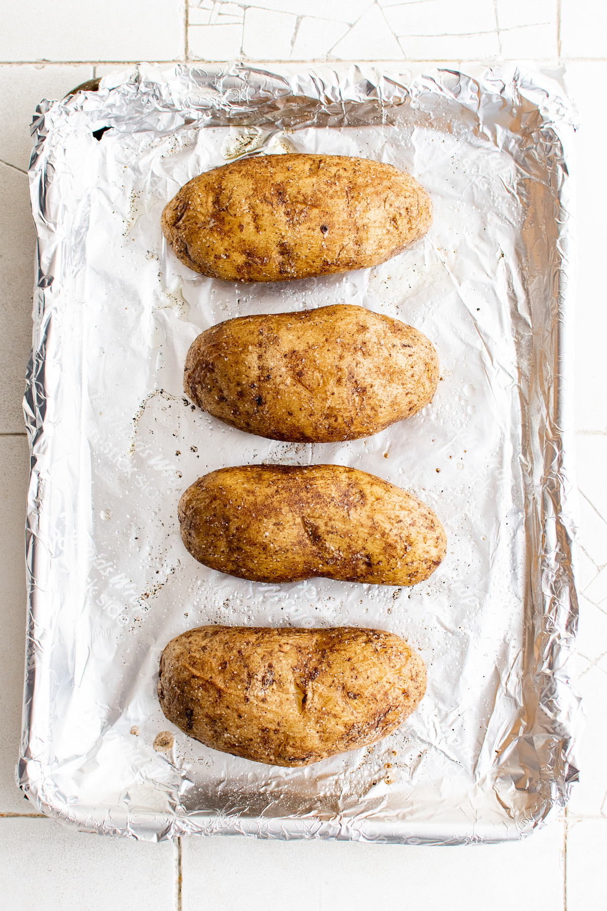Four potatoes that have been baked on a foil-lined baking sheet.