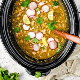 A crockpot filled with chicken pozole with lime wedges, cilantro and radishes on top.
