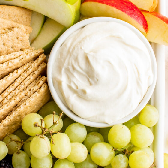 A bowl of creamy white dip surrounded by grapes, crackers, and apples.
