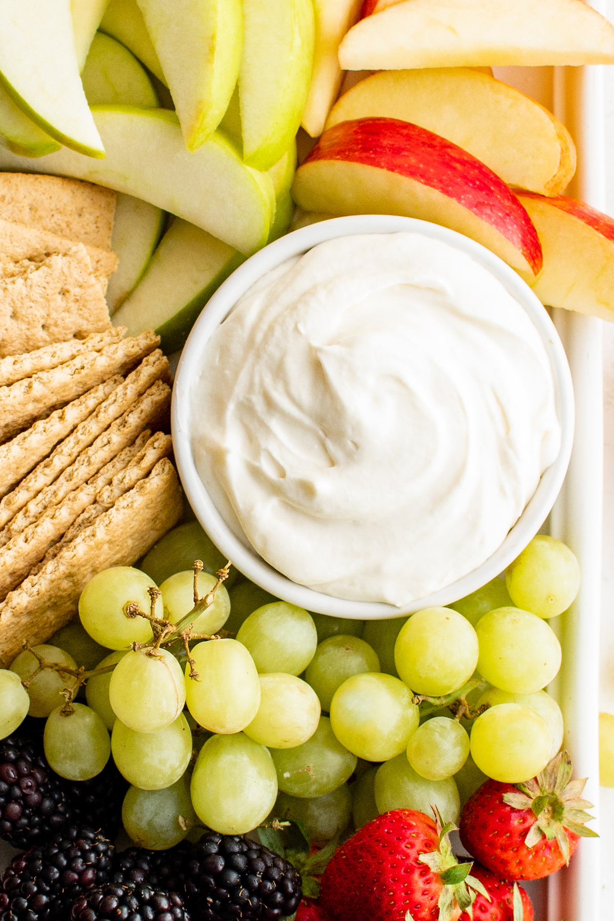 A bowl of creamy white dip surrounded by grapes, crackers, and apples.