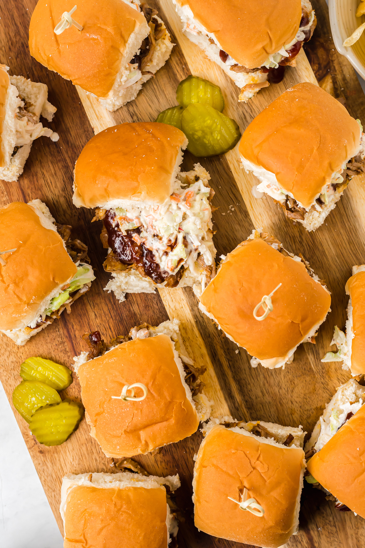 A dozen small barbecue sandwiches on a cutting board with pickles.
