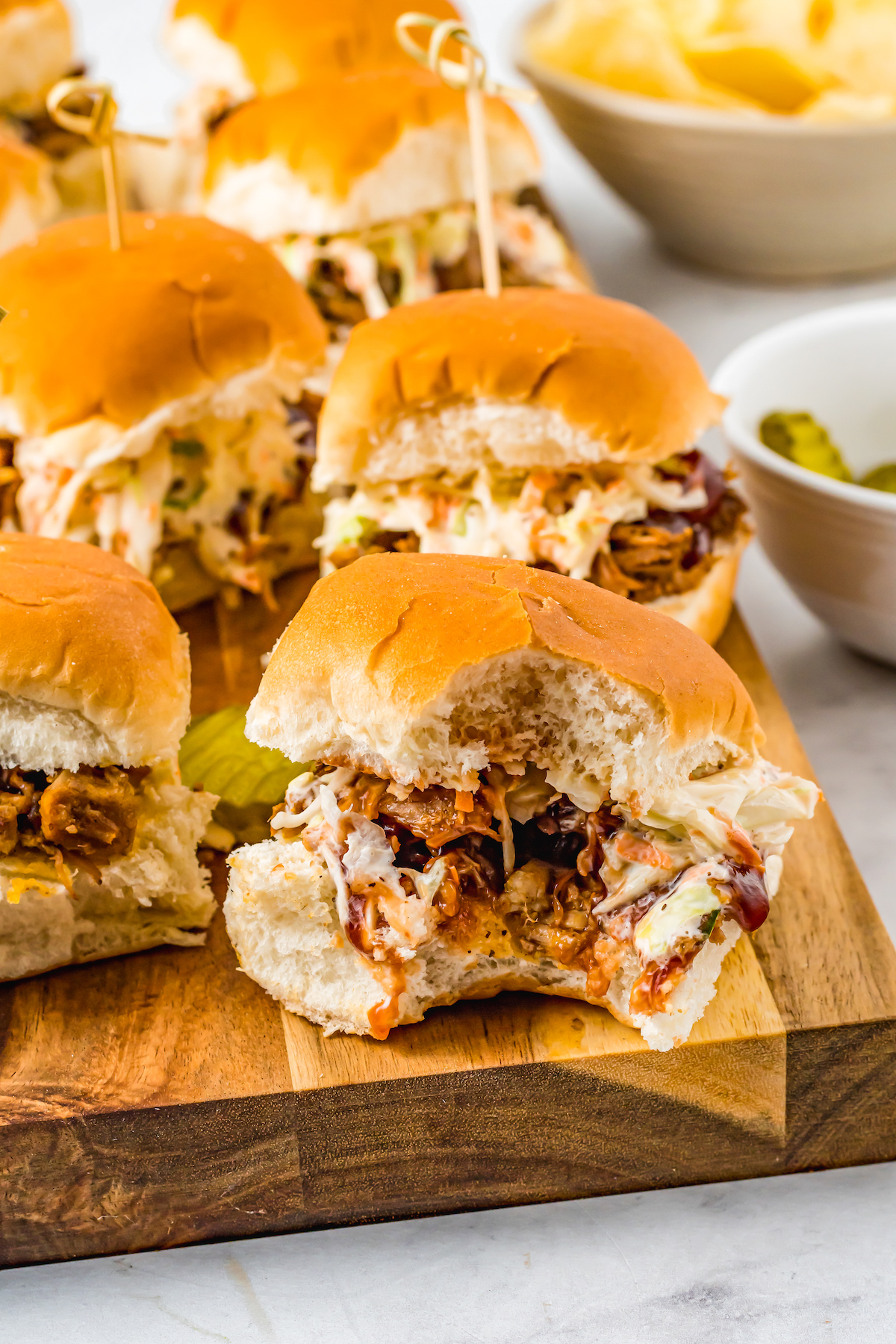 Pulled pork sliders on a cuttinng board. One has had a bite taken out of it.