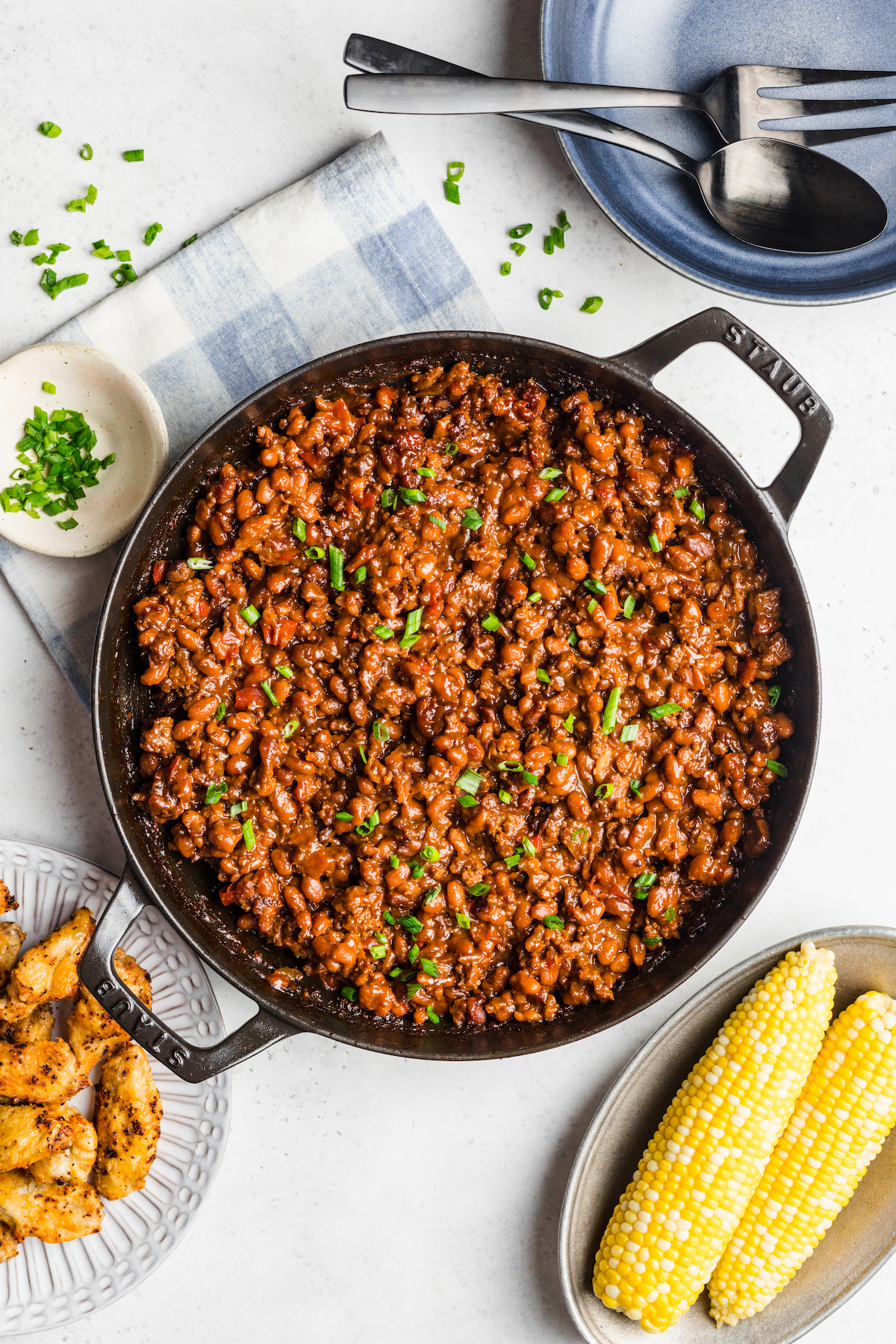 A double-handled skillet of baked beans on a table with ears of corn on the cob and other dishes.