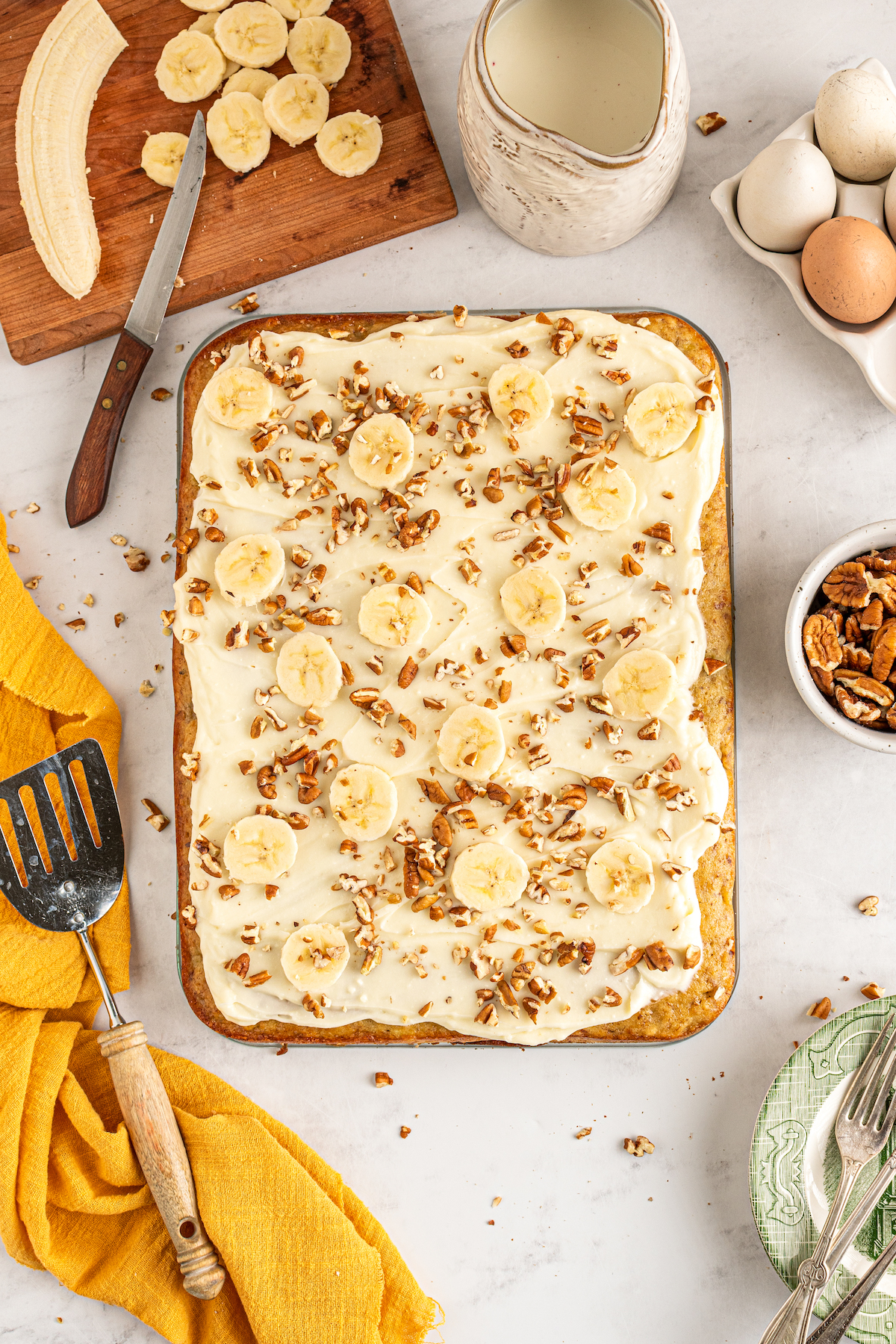 Banana bread cake on a table with sliced bananas, a bowl of pecans, a cloth napkin, and eggs.