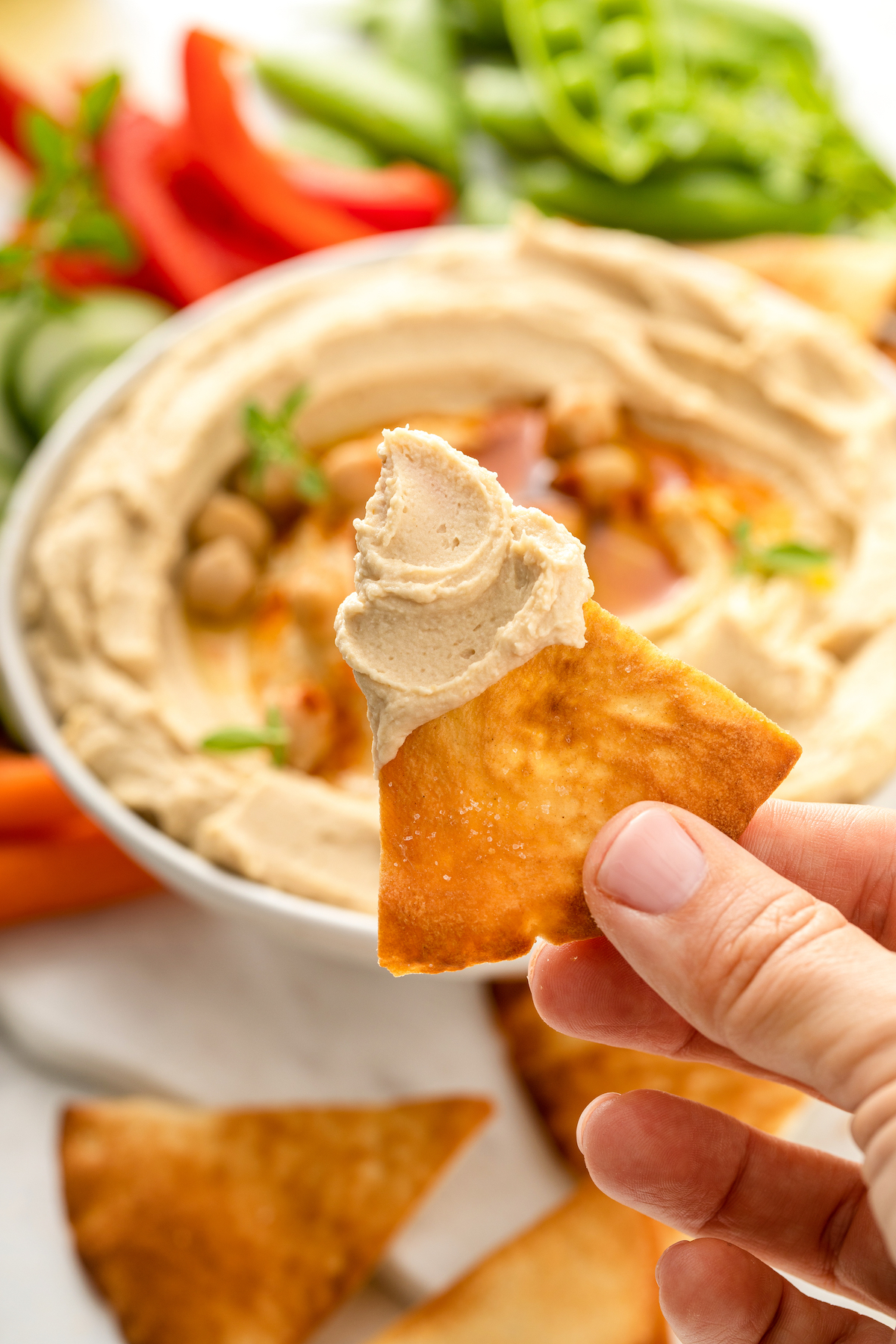 A pita chip dipped in chickpea dip and held toward the camera.