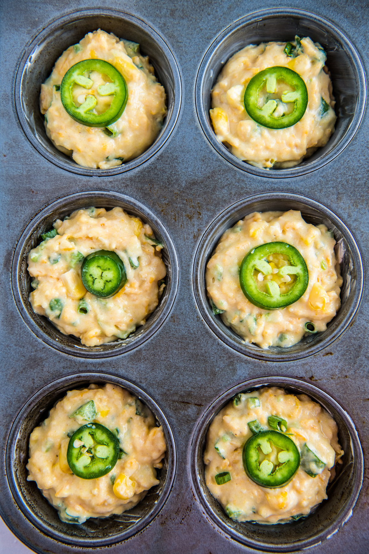 Overhead shot of a muffin tin. Each well is partially filled with batter and topped with a jalapeno slice.