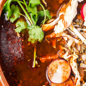 A large pot of pork pozole rojo with cilantro and radishes on top.