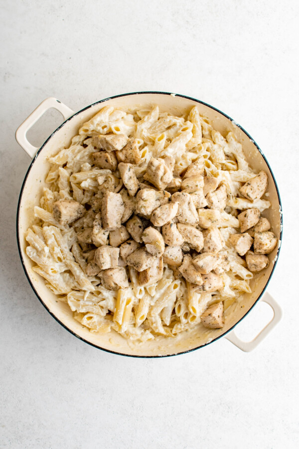 Pieces of cooked chicken, pasta, and alfredo sauce in a pan.