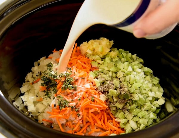 Evaporated milk being poured into a slow cooker filled with cut up vegetables.