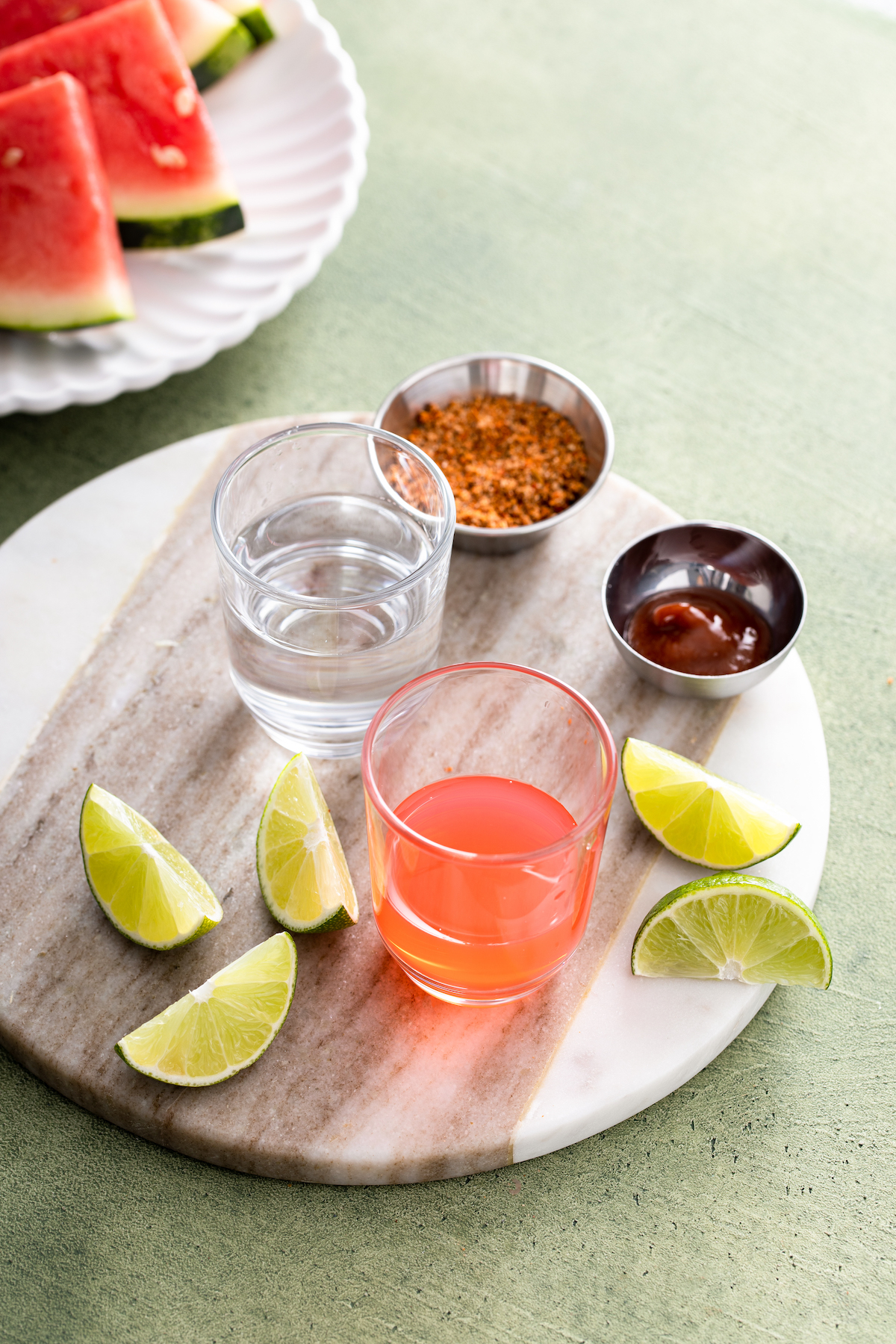 Tajin, hot sauce, lime wedges, watermelon schnapps, and tequila on a table.