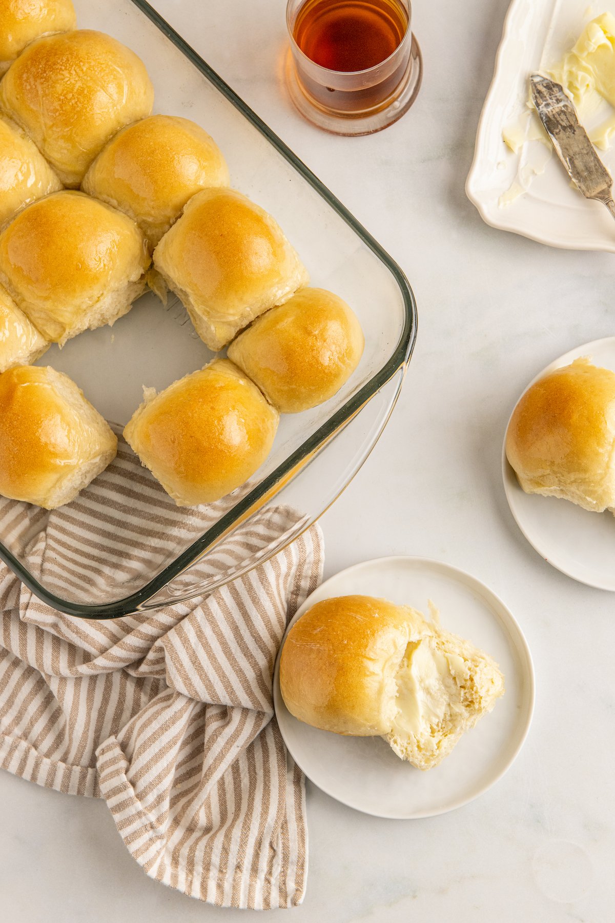 Overhead shot of a baking dish of rolls, with two rolls served onto small plates. A dish of butter and a dish of jelly sit nearby on the table.