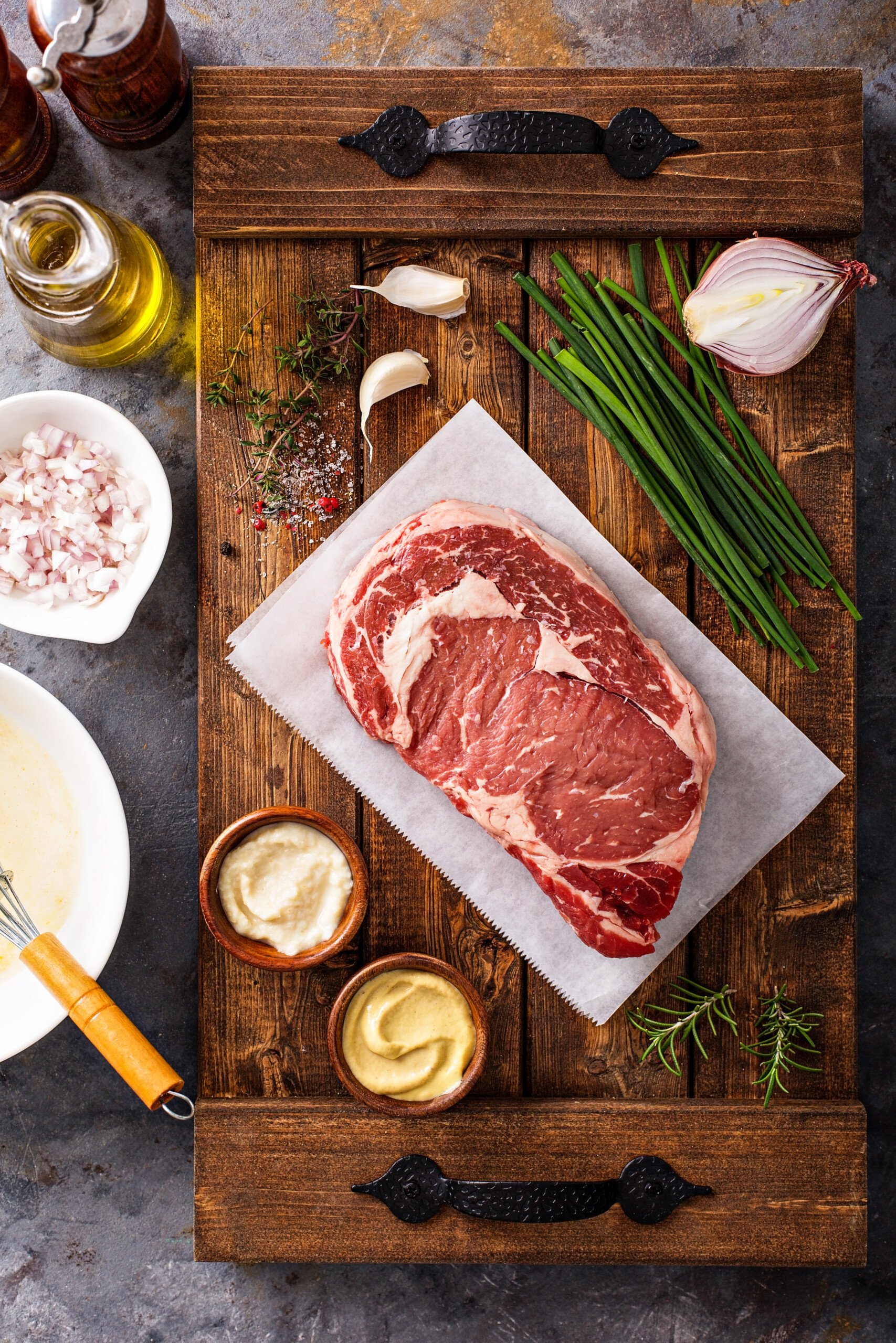 A steak on parchment paper surrounded by bowls of ingredients, fresh herbs and a shallot.