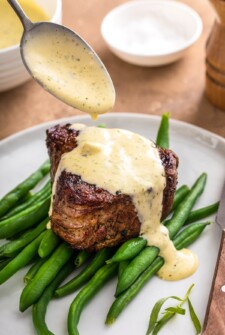 A spoon drizzling Béarnaise Sauce on top of a steak on a bed of green beans.
