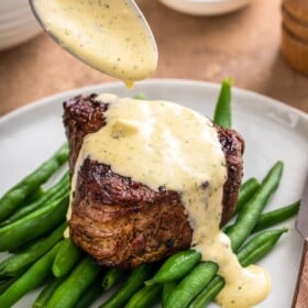 A spoon drizzling Béarnaise Sauce on top of a steak on a bed of green beans.