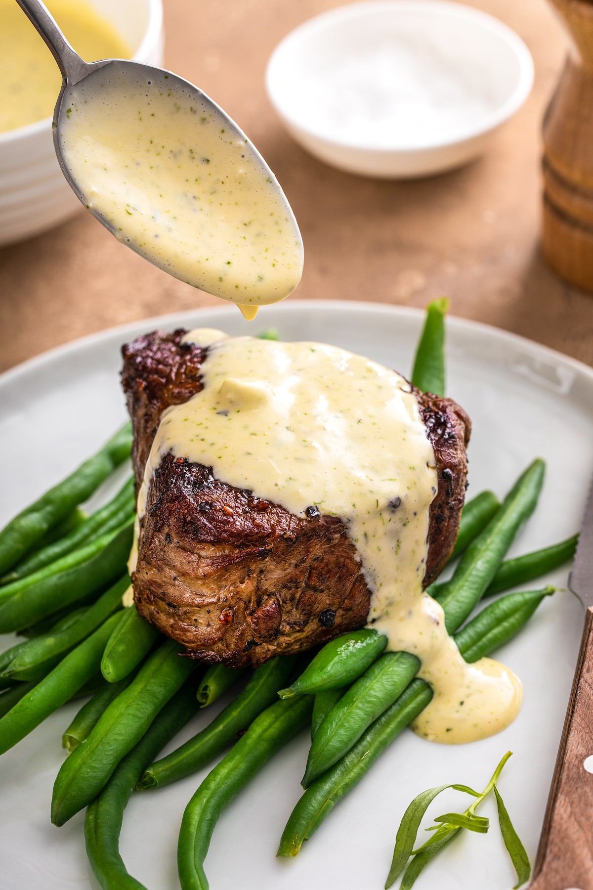 A spoon drizzling Béarnaise Sauce on top of a steak on a bed of green beans.
