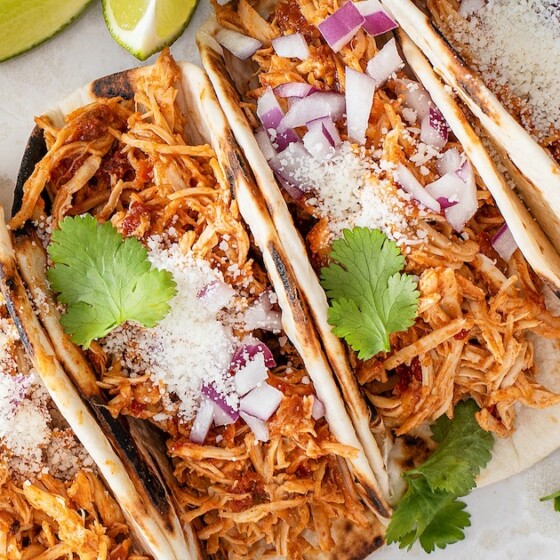 Crockpot chicken tacos on a platter with cilantro, cotija cheese, red onion and lime wedges.