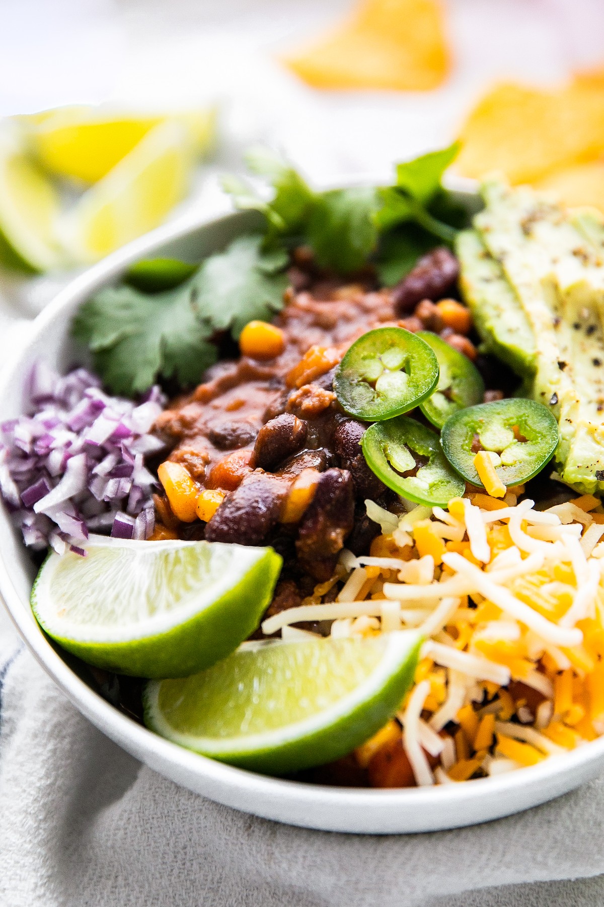 Chili in a white bowl with lime wedges, shredded cheese, and more.