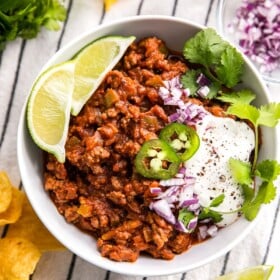 A bowl of chili topped with lime wedges, jalapenos, and onion.